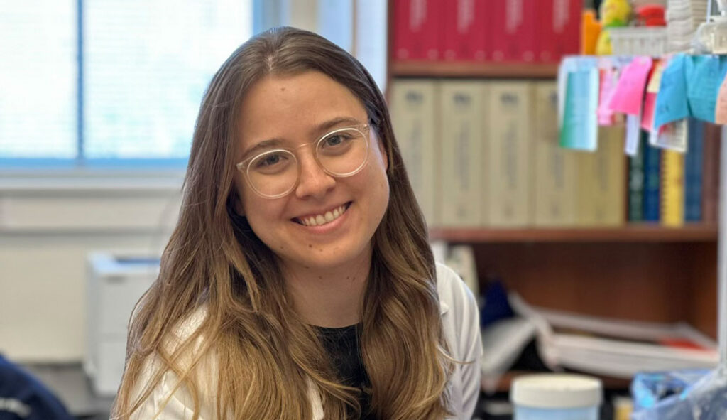 A graduate student with long brown hair and glasses, wearing a white lab coat, smiles in a laboratory setting focused on cellular biology, with shelves and books in the background.
