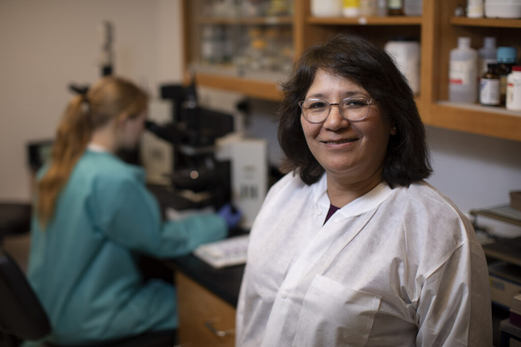 Two women in a laboratory; Ynes Ortega is in focus, wearing a white lab coat and glasses, smiling at the camera, while the other works at a microscope in the background—capturing a moment of scientific discovery worthy of the news.